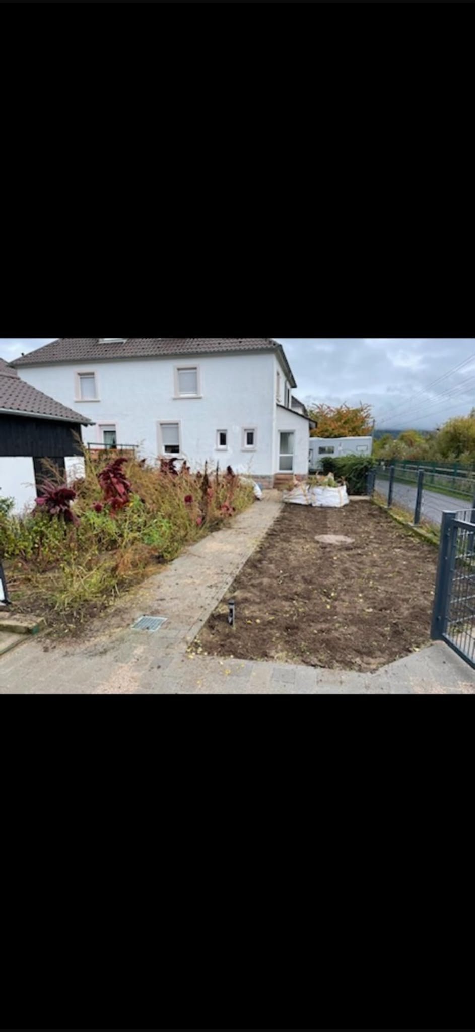 Zweigeschossiges weißes Haus mit dunklem Dach neben einer umgegrabenen Gartenfläche, die von einem Metallzaun eingefasst ist, davor herbstliche Vegetation