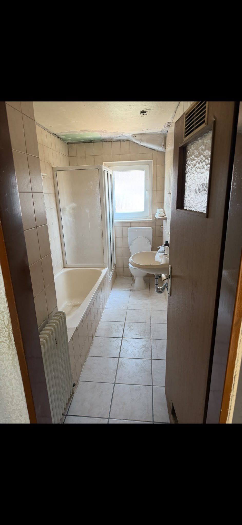 Schmales, fertiggestelltes Wannenbad mit beigen Fliesen, Duschabtrennung, Toilette und Handtuchhalter